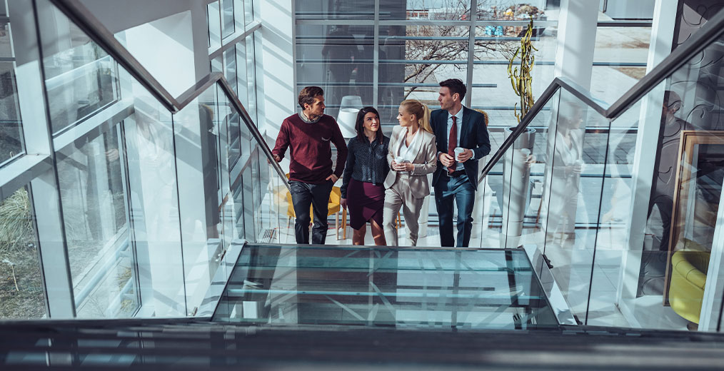 Vier Personen gehen gemeinsam eine gl&auml;serne Treppe hinunter und unterhalten sich in einem lichtdurchfluteten B&uuml;rogeb&auml;ude.