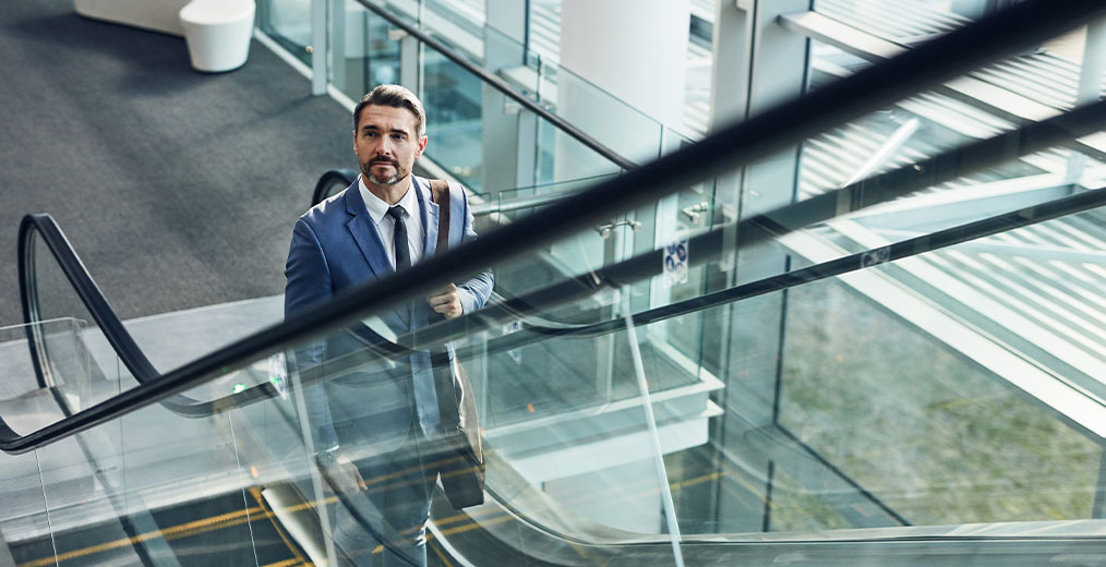 Ein Mann im Anzug f&auml;hrt mit einer Rolltreppe in einem lichtdurchfluteten B&uuml;rogeb&auml;ude nach oben und blickt konzentriert nach vorn.
