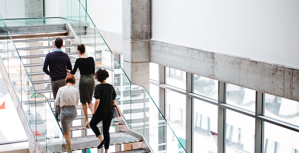 Vier Personen in Businesskleidung steigen eine gl&auml;serne Treppe in einem hellen, modernen B&uuml;rogeb&auml;ude hinauf.