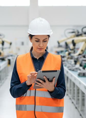 Ingenieurin mit Schutzhelm und Sicherheitsweste arbeitet mit Tablet in einer modernen Produktionshalle.