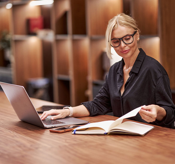 Eine Frau mit Brille sitzt an einem Schreibtisch, arbeitet am Laptop und pr&uuml;ft gleichzeitig Unterlagen &ndash; Symbol f&uuml;r den Rechts- und Compliance-Bereich.