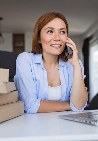 L&auml;chelnde Frau f&uuml;hrt ein Telefonat am Laptop, neben ihr liegen mehrere Kartons.