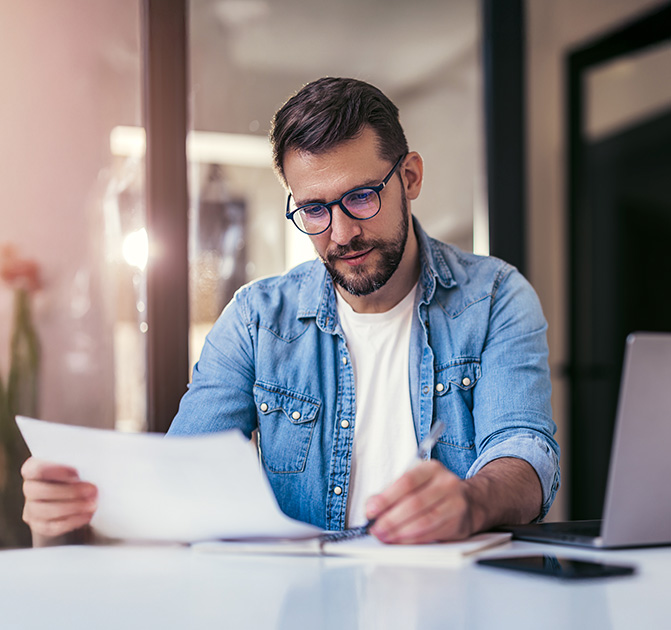 Nachdenklicher Mann mit Brille schreibt und pr&uuml;ft Unterlagen am Schreibtisch.