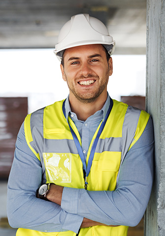 Ein Bauleiter tr&auml;gt Schutzhelm und Warnweste und l&auml;chelt selbstbewusst auf einer Baustelle.