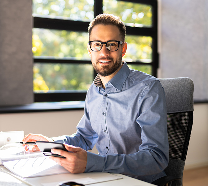 Gesch&auml;ftsmann mit Brille sitzt am Schreibtisch und arbeitet mit Tablet und Unterlagen.
