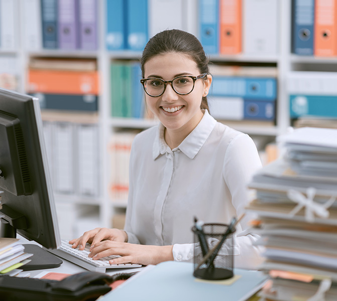 Eine junge Frau mit Brille sitzt an einem Schreibtisch im B&uuml;ro, tippt auf einer Tastatur und l&auml;chelt freundlich in die Kamera. Im Hintergrund sind bunte Aktenordner in Regalen zu sehen.