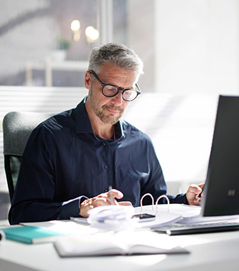 Ein &auml;lterer Mann mit Brille arbeitet konzentriert an seinem Schreibtisch, vor ihm liegen Unterlagen und ein Ordner. Er sitzt zwischen zwei gro&szlig;en Monitoren in einem hellen B&uuml;ro mit viel Tageslicht.