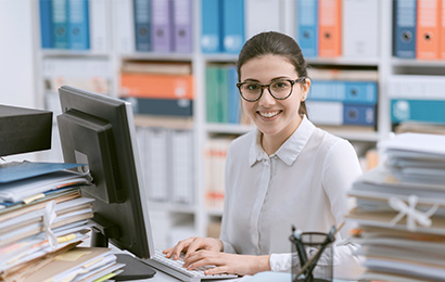 Junge Frau arbeitet am PC im B&uuml;ro und l&auml;chelt in die Kamera.