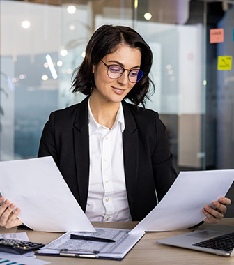 Frau im B&uuml;ro mit Laptop und Papieren &uuml;berpr&uuml;ft Unterlagen.