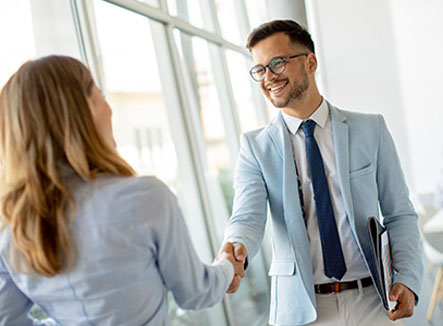 Zwei Personen begr&uuml;&szlig;en sich mit einem Handschlag in einem hellen B&uuml;ro, im Hintergrund arbeitet eine weitere Person.