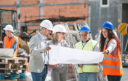 Vier Personen mit Schutzhelmen betrachten gemeinsam Baupl&auml;ne auf einer Baustelle.