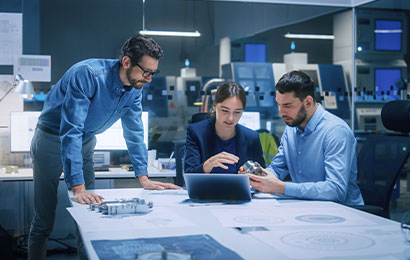 Drei Personen besprechen technische Zeichnungen und Bauteile an einem Tisch im B&uuml;ro.