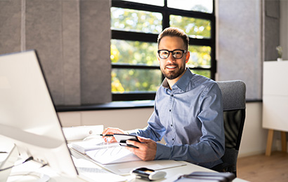 esch&auml;ftsmann mit Brille arbeitet l&auml;chelnd am Schreibtisch mit Laptop und Unterlagen.