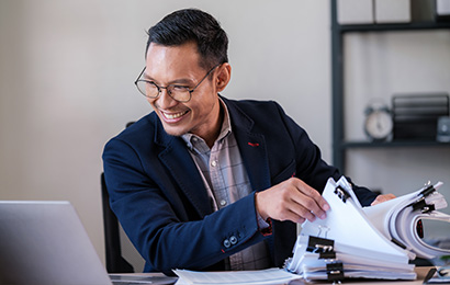 Gesch&auml;ftsmann mit Brille sortiert Unterlagen am Schreibtisch und lacht.