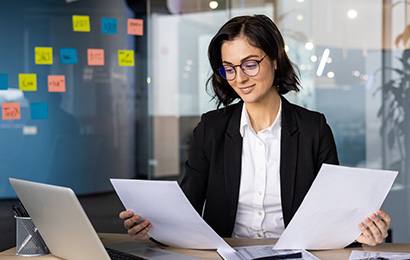 Gesch&auml;ftsfrau mit Brille pr&uuml;ft Unterlagen am Schreibtisch mit Laptop und Taschenrechner.