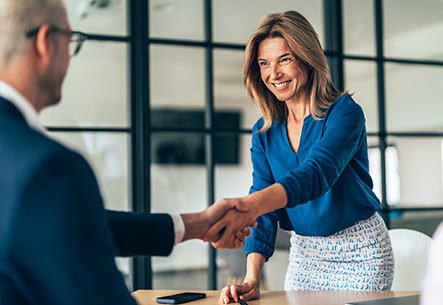 Eine l&auml;chelnde Frau in blauer Bluse sch&uuml;ttelt im B&uuml;ro einem gegen&uuml;berstehenden Mann die Hand.