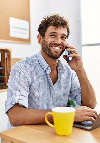 Gut gelaunter Mann spricht am Smartphone und sitzt an einem Schreibtisch mit Laptop.