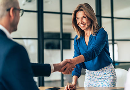 L&auml;chelnde Frau in blauer Bluse sch&uuml;ttelt einem Kollegen im Besprechungsraum die Hand.