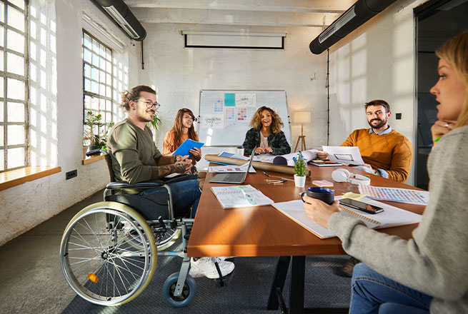 Eine Gruppe von f&uuml;nf Personen, darunter eine Person im Rollstuhl, arbeitet gemeinsam an einem Tisch.