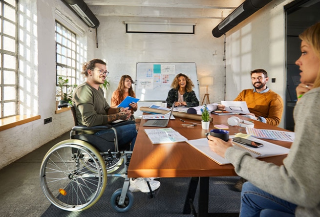Eine Gruppe von f&uuml;nf Personen, darunter eine Person im Rollstuhl, arbeitet gemeinsam an einem Tisch.