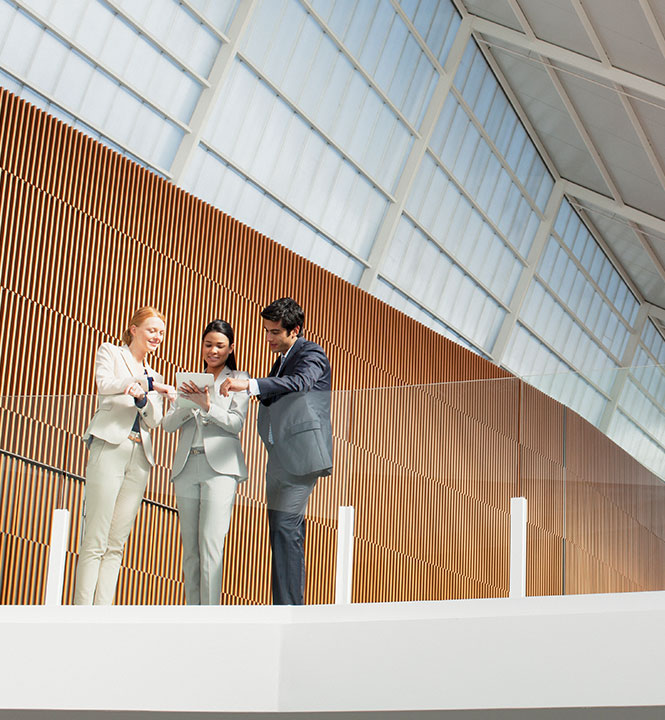 Drei Personen im Business-Outfit stehen in einem modernen Geb&auml;ude und sehen sich ein Tablet an.