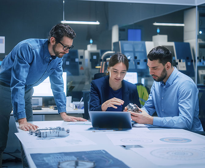 Drei Personen besprechen technische Zeichnungen und Bauteile an einem Tisch im B&uuml;ro.