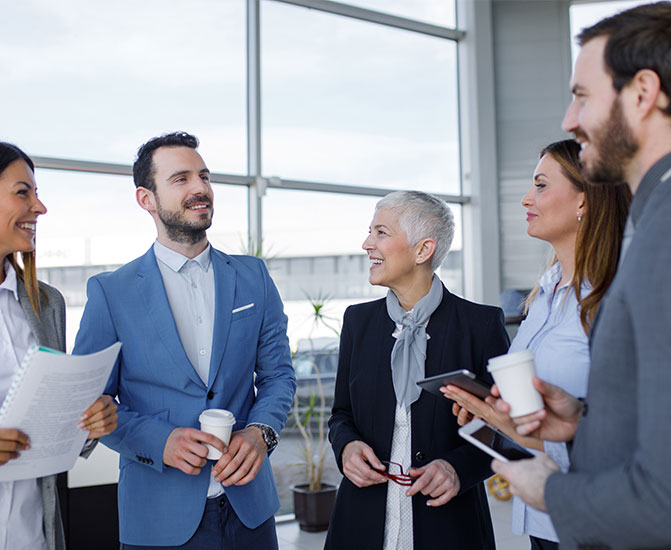 Vier Kolleginnen und Kollegen unterhalten sich in einem hellen B&uuml;rogeb&auml;ude und l&auml;cheln &ndash; Symbol f&uuml;r erfolgreiche Zusammenarbeit und Kommunikation im Team.