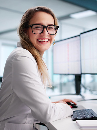 Analystin l&auml;chelt am Arbeitsplatz vor mehreren Monitoren mit Daten- und Tabellenansichten.