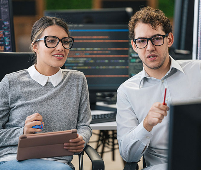 Zwei IT-Fachkr&auml;fte sitzen gemeinsam vor Monitoren mit Code und besprechen Programmierl&ouml;sungen.