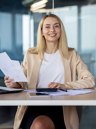 Frau sitzt am Schreibtisch, h&auml;lt Dokumente in der Hand und l&auml;chelt freundlich.