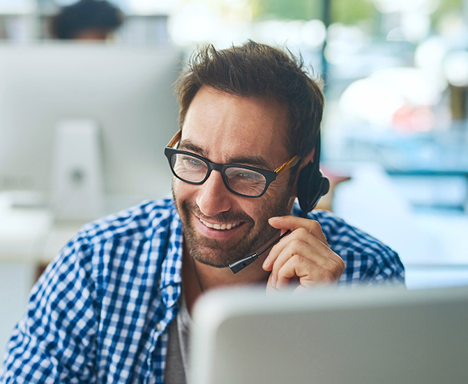 Ein Mann mit Brille und kariertem Hemd sitzt in einem modernen B&uuml;ro, tr&auml;gt ein Headset und schaut l&auml;chelnd auf den Bildschirm, w&auml;hrend er offenbar ein Gespr&auml;ch f&uuml;hrt oder jemandem am Computer hilft.
