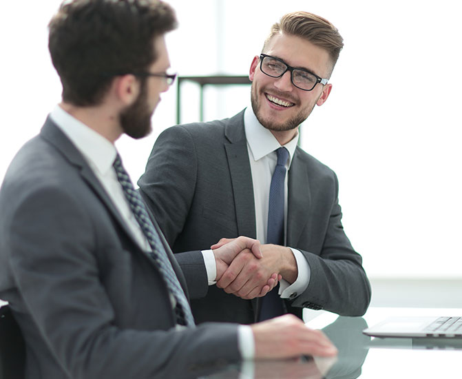 Zwei M&auml;nner im Anzug sitzen an einem Tisch und begr&uuml;&szlig;en sich mit einem freundlichen H&auml;ndedruck.