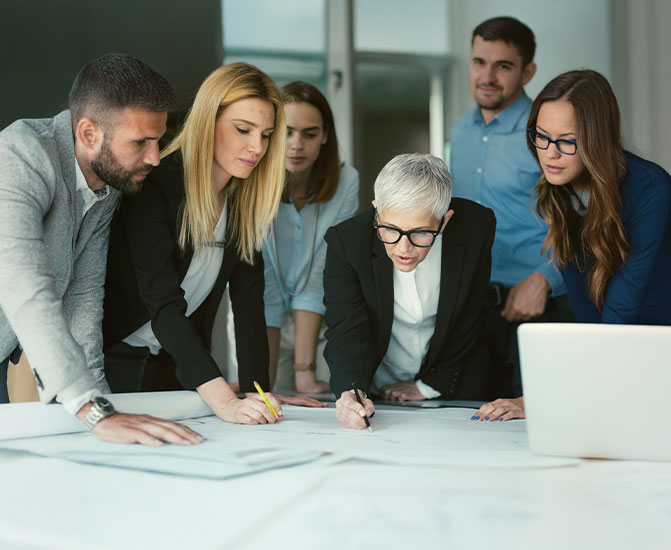 Sechs Personen stehen um einen Tisch und analysieren gemeinsam Dokumente und Pl&auml;ne.