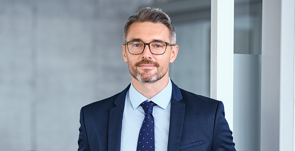 Selbstbewusster Mann in Anzug und Krawatte steht in modernem B&uuml;ro und blickt in die Kamera.