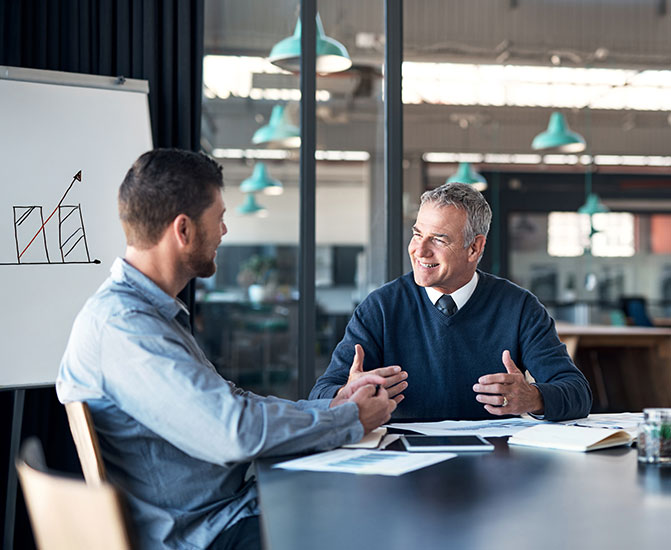 Zwei M&auml;nner sitzen an einem Konferenztisch und f&uuml;hren ein positives, fachliches Gespr&auml;ch, w&auml;hrend im Hintergrund eine Grafik mit wachsendem Diagramm zu sehen ist &ndash; Symbol f&uuml;r Unternehmenswachstum und Zusammenarbeit.